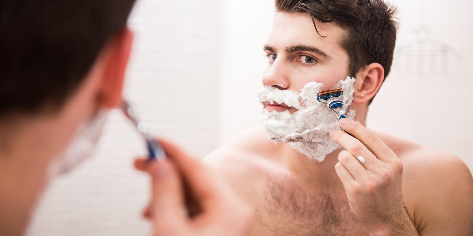Handsome young man is shaving his face and looking at the mirror.Shaving with fun. Rear view of playful young man with shaving cream on his face is standing in front of the mirror and smiling.Young handsome man holds pepper, isolated on white background.