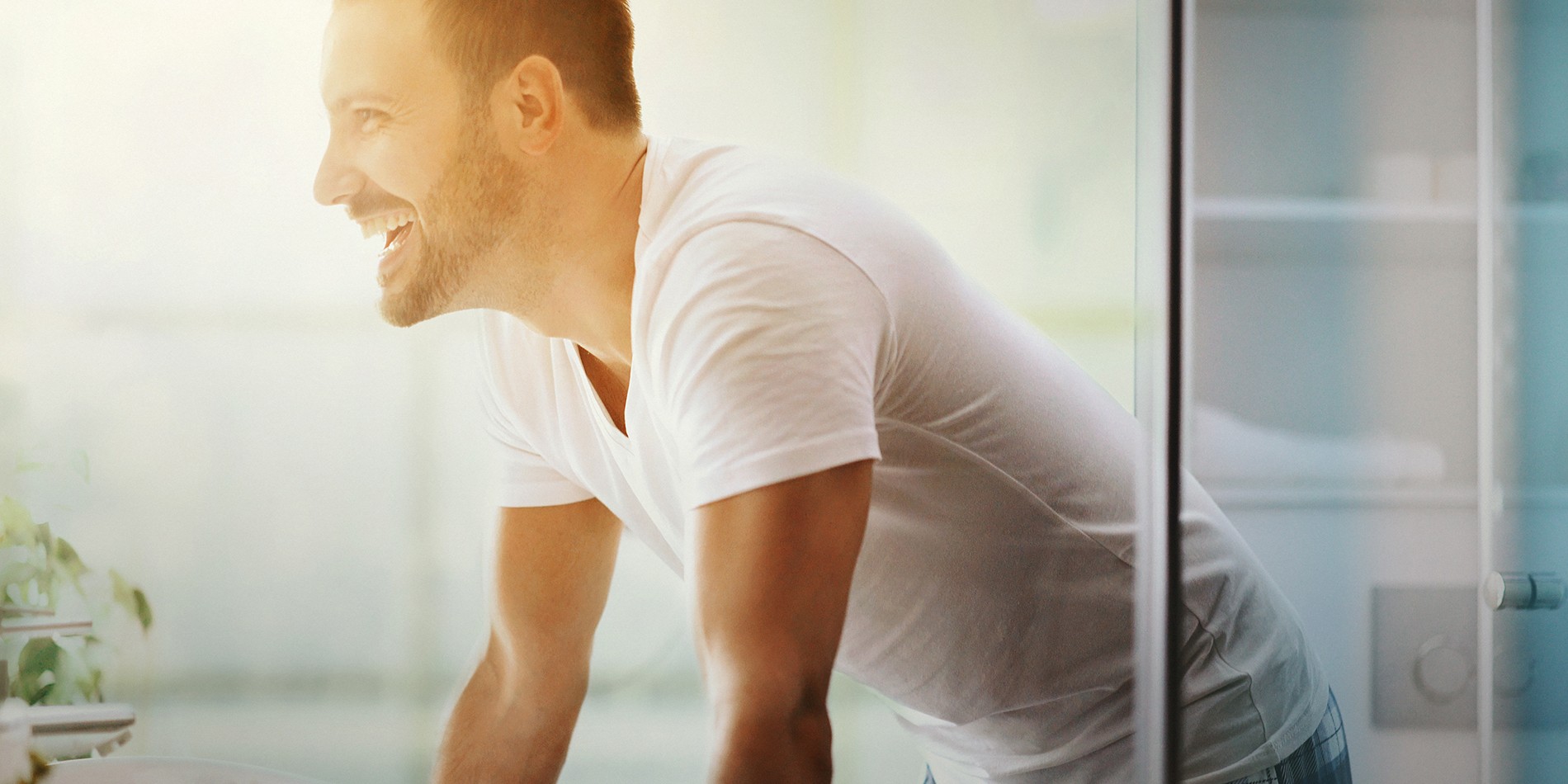 Closeup side view of a cheerful late 20's man leaning over bathroom sink and looking at his reflection in the mirror. He's lit by warm and low morning light coming through the window in the back.