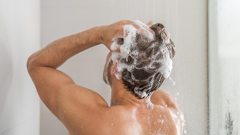 Man wast zijn haren met schuimende shampoo onder de douche.						 Man wast zijn haren met schuimende shampoo onder de douche.
